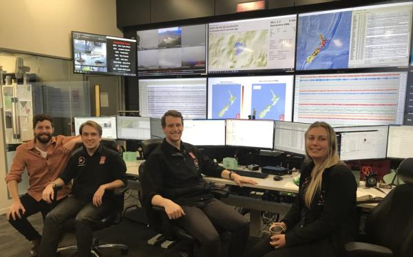 Four people on office chairs smile at the camera while sitting in front of an array of six computer mo<em></em>nitors and ten large screens showing maps and data.