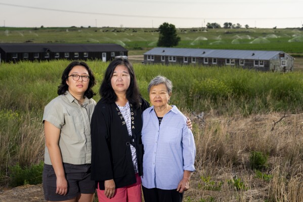 Minidoka survivor Mary Tanaka Abo, 83, at right, poses for a portrait with daughter Julie Abo, center, and granddaughter Maya Abo Dominguez, left, in front of a historic mess hall and barracks at the Minidoka Natio<em></em>nal Historic Site, Saturday, July 9, 2023, in Jerome, Idaho. Tanaka Abo says she didn't want Julie or Maya to feel the same sense of shame surrounding incarceration as she did. Instead, she chose to speak openly a<em></em>bout her experience with her daughter and granddaughter, and has attended multiple pilgrimages with her family. (AP Photo/Lindsey Wasson)