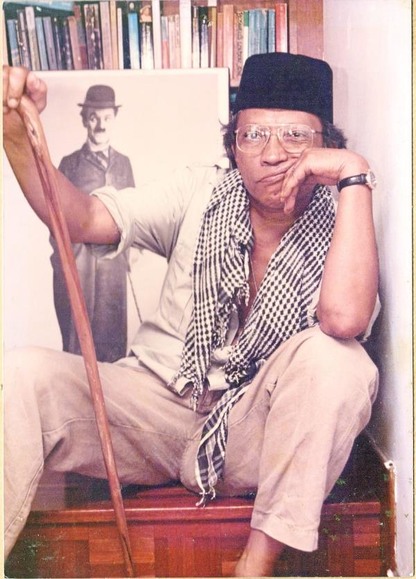 Salleh Ben Jo<em></em>ned in a so<em></em>ngkok and cane, posing in front of a photo of Charlie Chaplin. Photo: Salleh Ben Jo<em></em>ned Literary Legacy Project